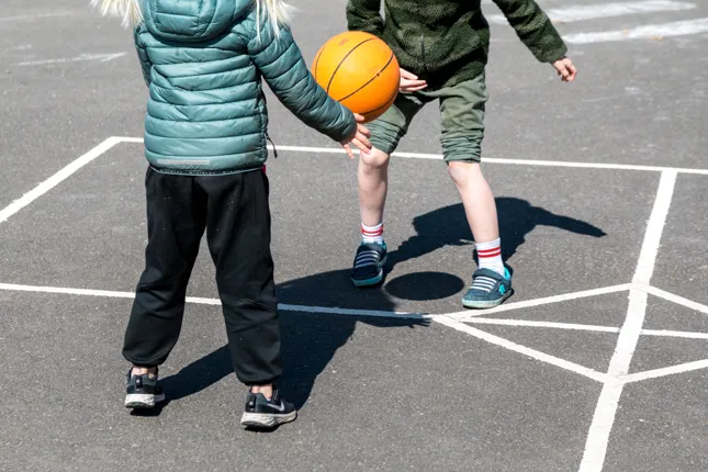 Två barn leker med en basketboll på en parkeringsplats.